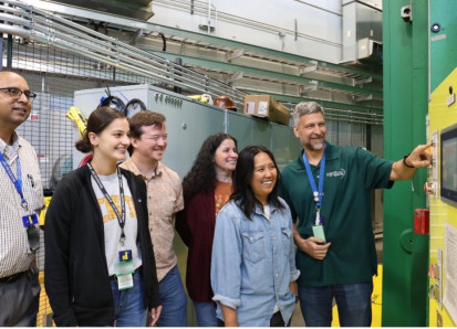 A group of six researchers in a lab stand beside industrial equipment, smiling as one man operates a