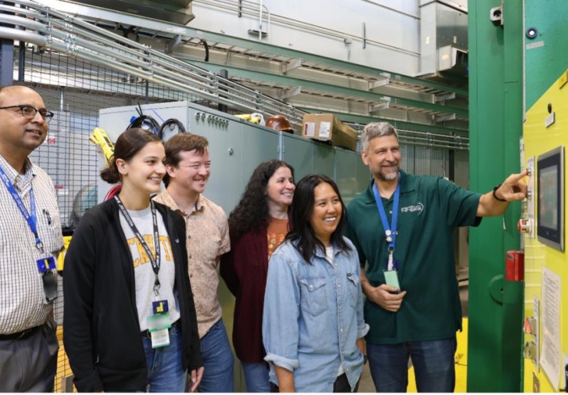 A group of six researchers in a lab stand beside industrial equipment, smiling as one man operates a