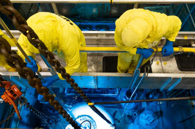 Two reactor operators wearing yellow protective suits and blue gloves work from a bridge platform ab