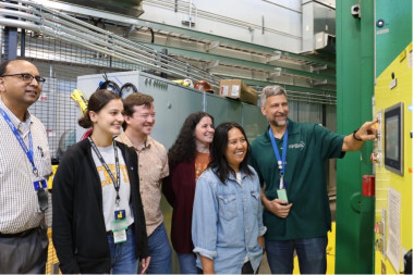 A group of six researchers in a lab stand beside industrial equipment, smiling as one man operates a