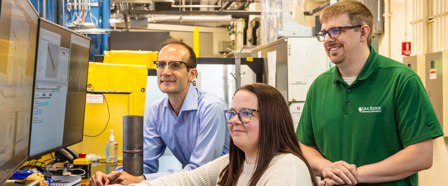 Three scientists looking at monitor. Two are standing and one is sitting in a chair